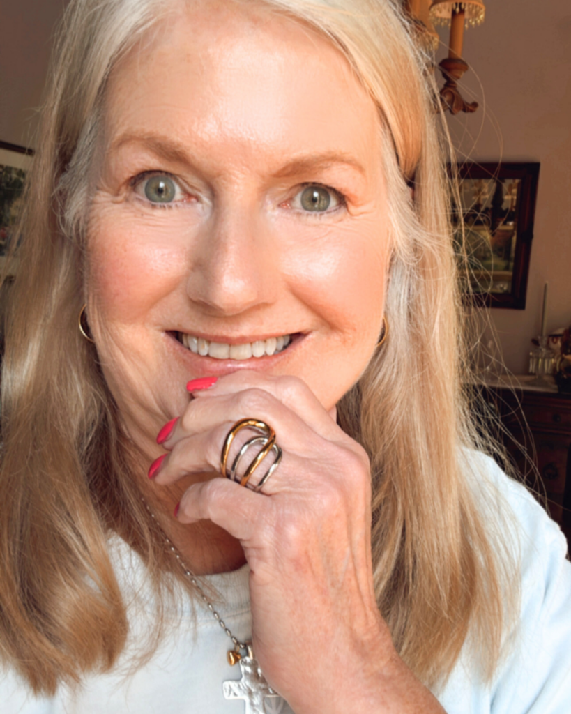 Smiling woman with long blonde hair wearing a white top and cross necklace, showing off a bold two-tone stainless and gold statement ring on her hand with bright pink nail polish.