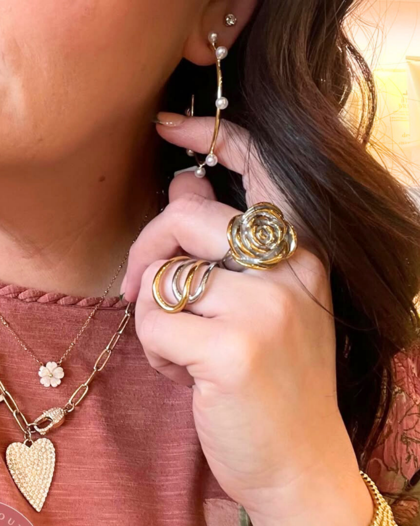 Close-up of a woman wearing gold and pearl hoop earrings, layered gold necklaces, and two bold rings—one rose-shaped and one two-tone swirl design—styled on her hand near her face. She is dressed in a mauve top with a floral outer layer.