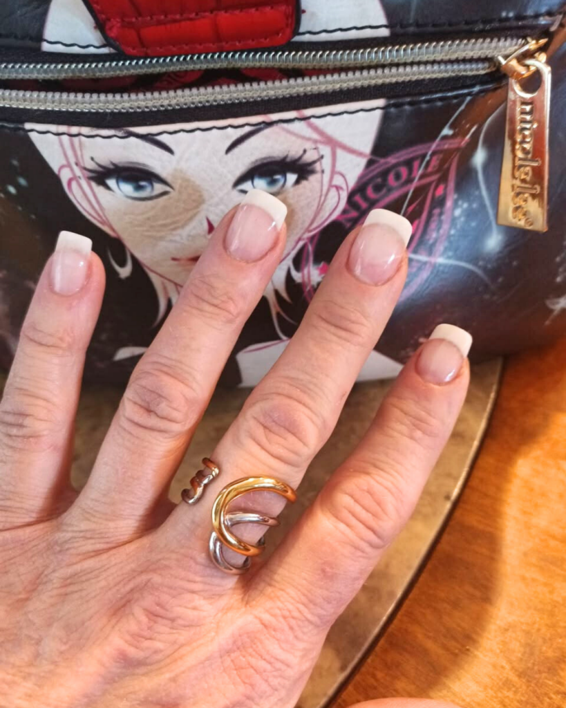 Close-up of a hand with French manicured nails wearing two rings—one rose gold open-style ring and one bold two-tone stainless and gold swirl ring—posed in front of a designer bag featuring illustrated artwork and a metallic zipper pull.