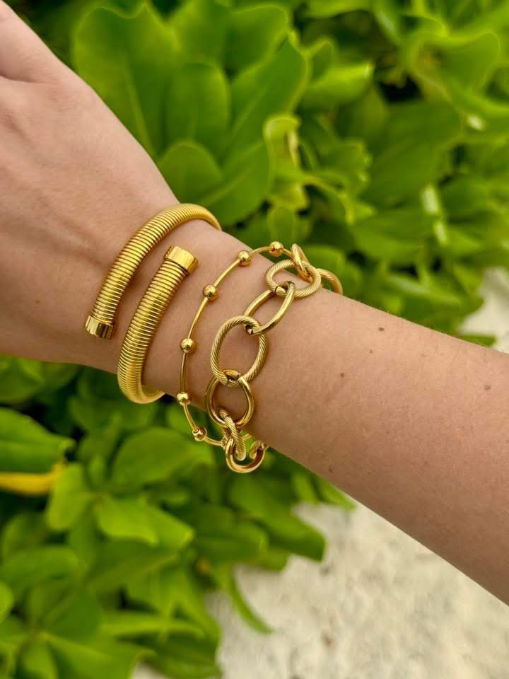Woman’s wrist with a gold Tamia Cuff Bracelet stacked with gold chains and beaded bracelets in front of green foliage.