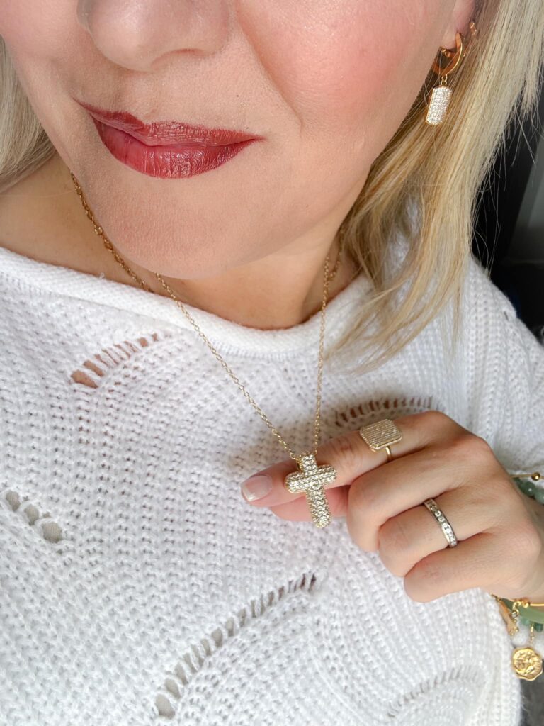 Close-up of a woman wearing a gold crystal cross necklace, gold crystal drop earrings, a textured white knit top, and coordinating gold rings and bracelets, showcasing a faith-inspired jewelry look.