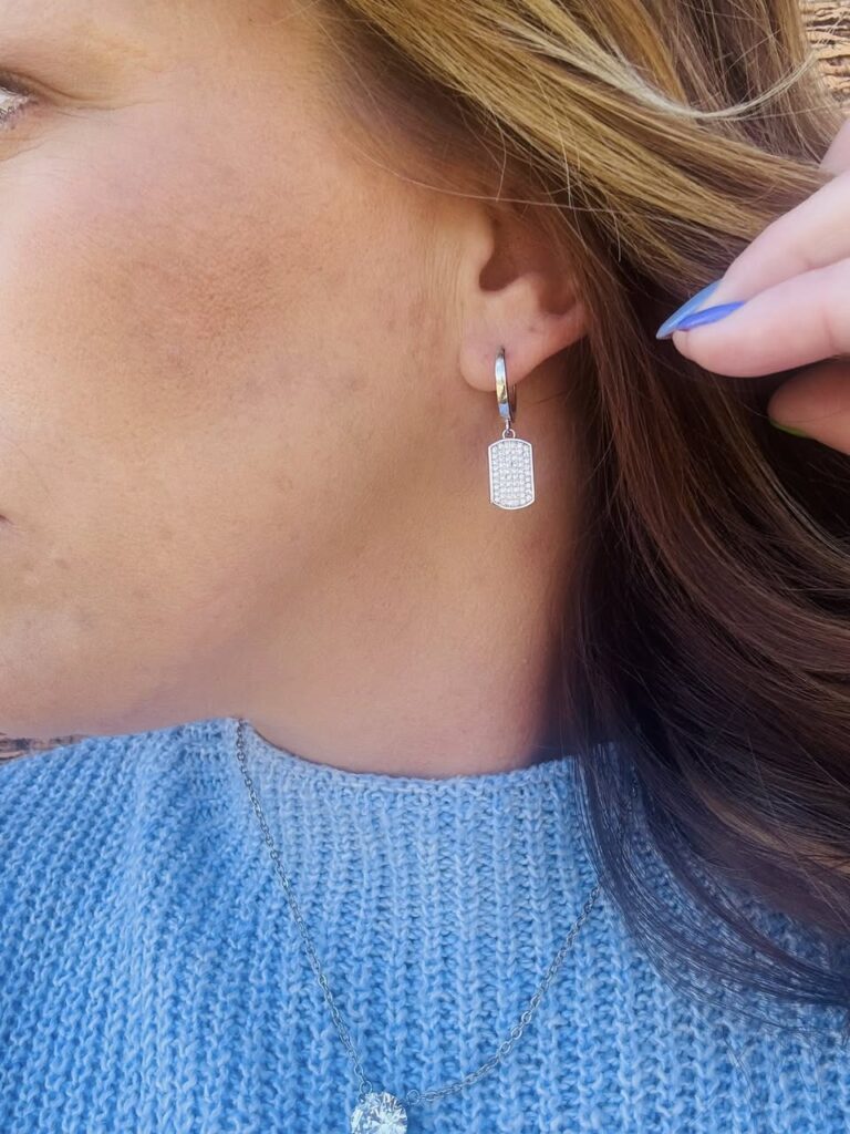 Close-up of a woman wearing silver hoop earrings with a rectangular crystal drop and a delicate silver chain necklace with a round pendant, styled with a light blue knit sweater.