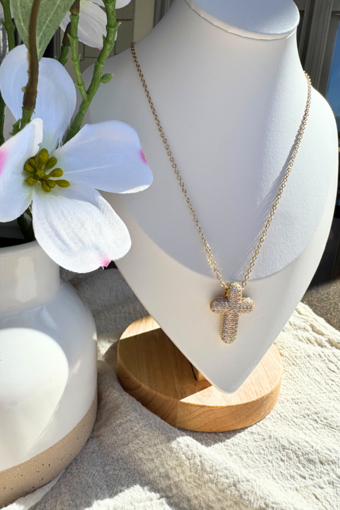 Gold crystal cross necklace displayed on a white jewelry stand with a wooden base, styled beside a white ceramic vase with artificial white flowers on a neutral fabric background.
