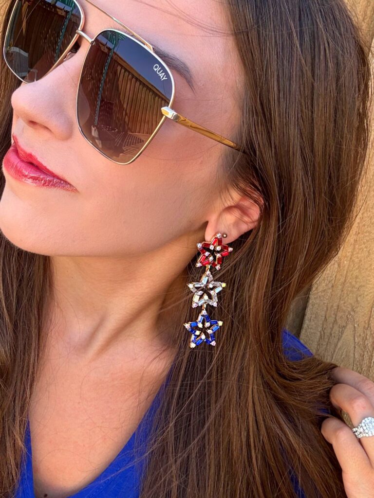Close-up of a woman wearing oversized gold-rimmed Quay sunglasses and bold, dangling star earrings in red, white, and blue. She is wearing a royal blue top and has long brown hair. The earrings feature three stacked stars with rhinestone embellishments, perfect for patriotic or Fourth of July styling.