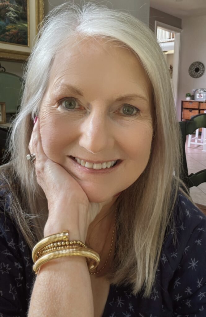 Smiling woman with long silver hair resting her chin on her hand, wearing a navy top with star patterns, a gold bracelet stack, and a wedding ring. She is sitting indoors with warm lighting and a cozy background featuring framed art and a wicker cabinet.