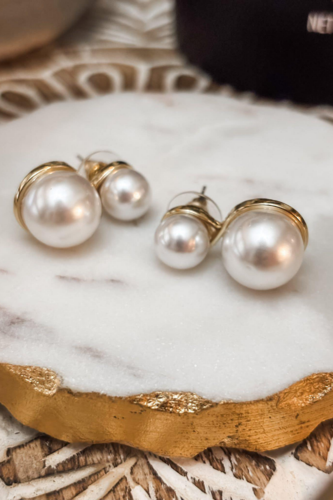 Close-up of gold double-tier pearl stud earrings resting on a round marble dish with a gold-trimmed edge. The JBloom Elizabeth Earrings are displayed to highlight their elegant, layered faux pearl design.