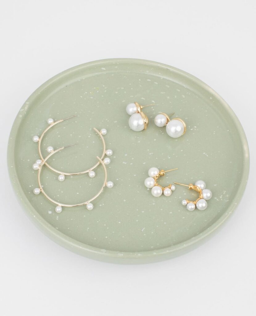 Flat lay of three pairs of modern pearl earrings on a sage green ceramic dish. Includes oversized double pearl studs, gold semi-hoops with spaced pearls, and curved mini hoops with clustered pearls—elegant and lightweight jewelry perfect for everyday or special occasions.