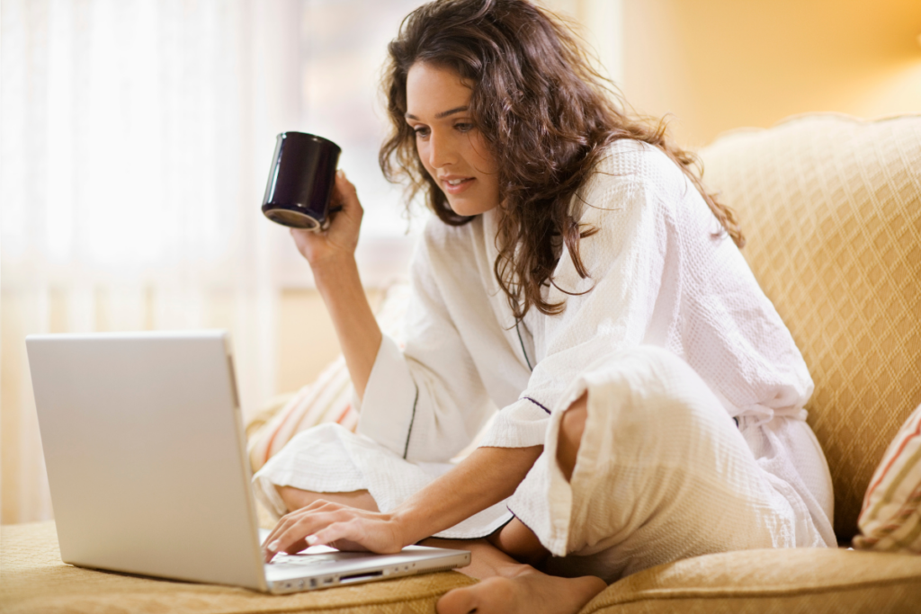 Woman in a white robe sitting on a couch with a mug in one hand and using a laptop on her lap, appearing relaxed and focused in a cozy home setting.
