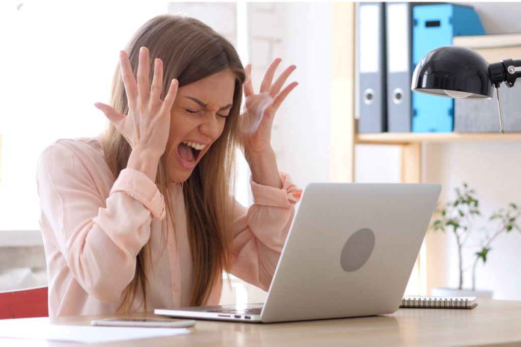 Frustrated woman yelling at her laptop with hands raised in exasperation while sitting at a desk in a modern office setting.