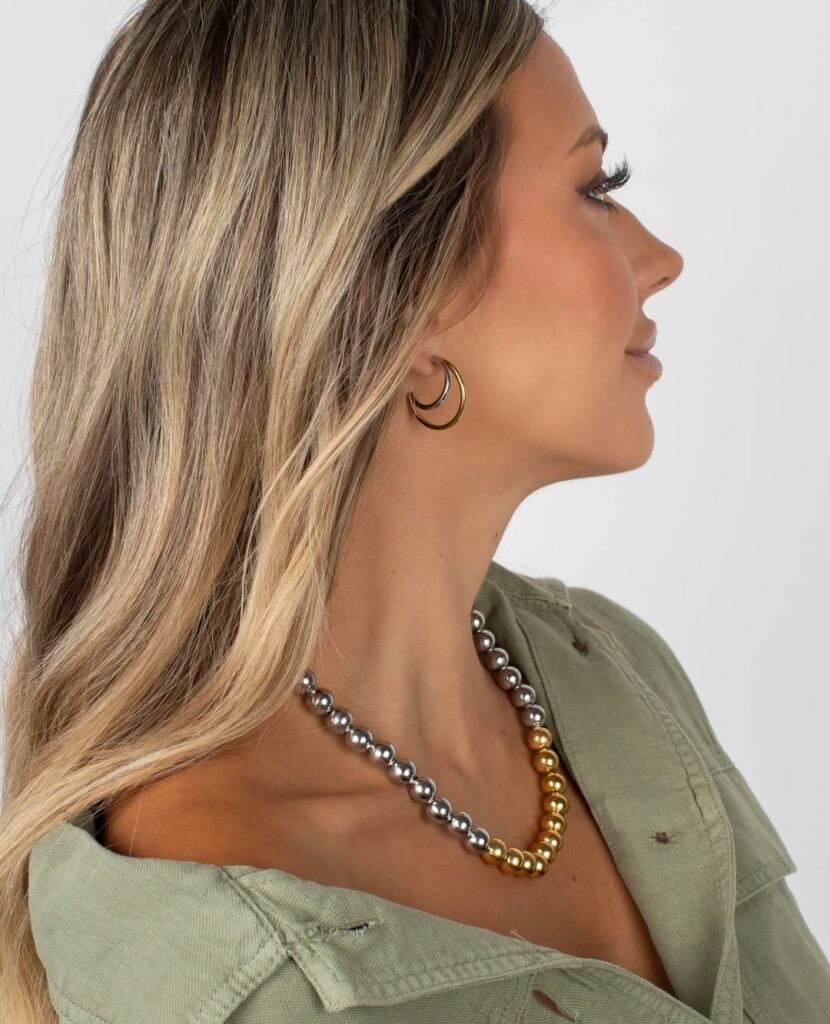 A woman wearing the Oaklyn Necklace, a stainless and gold stainless bubble bead necklace, paired with the Cabi Earrings, two-tone double hoop earrings. The jewelry blends silver and gold tones for a chic, versatile look, perfect for everyday style.