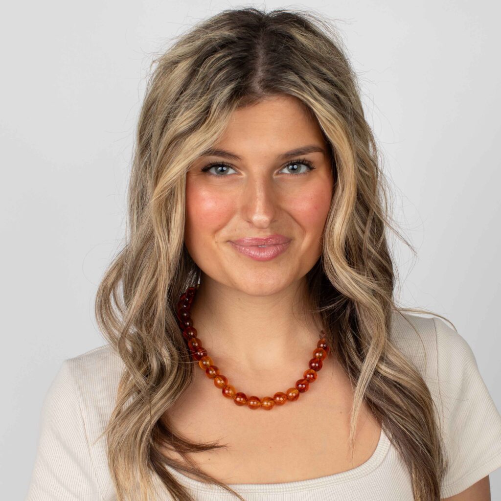 Woman wearing a burnt-orange marbled resin bead necklace with a cream scoop-neck top, smiling against a light gray background.