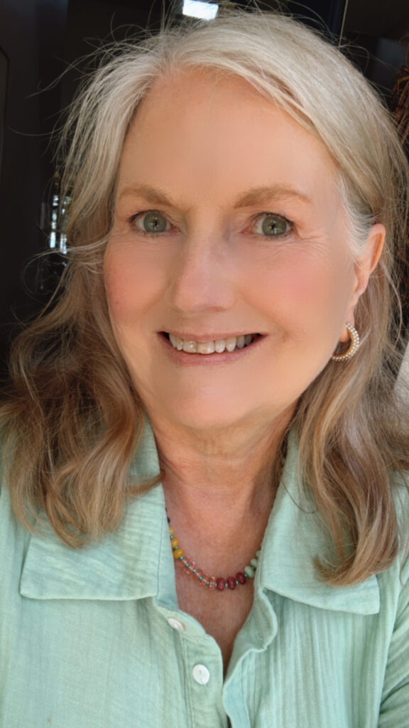 Smiling woman with long light brown hair wearing gold hoop earrings adorned with small pearls, a pastel green button-up shirt, and a colorful beaded necklace, photographed in natural light.